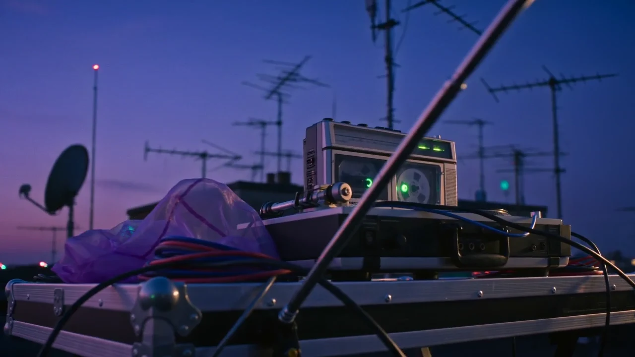 A sweeping rooftop camera move past a spinning cassette deck and city antennas